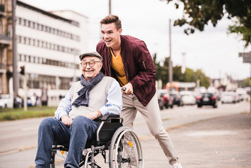 un jeune homme marche dehors avec un homme plus âgé assis dans un fauteuil roulant